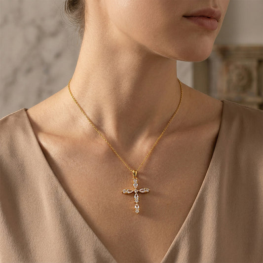 A close-up of a woman wearing a delicate gold cross pendant necklace adorned with small clear stones, paired with a beige satin top.