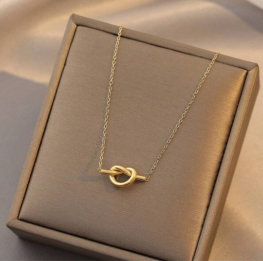 A gold necklace with a knot pendant displayed on a textured taupe jewelry box.
