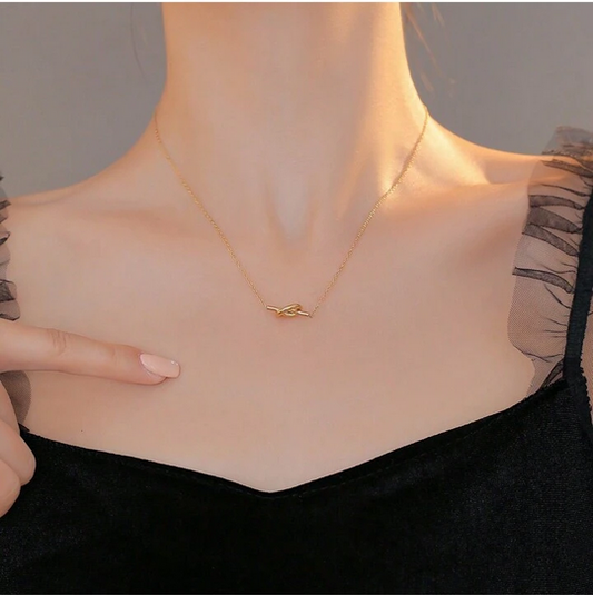 A close-up of a model's neck and chest wearing a delicate gold chain necklace with a small abstract wave or leaf-shaped pendant, paired with a black top.
