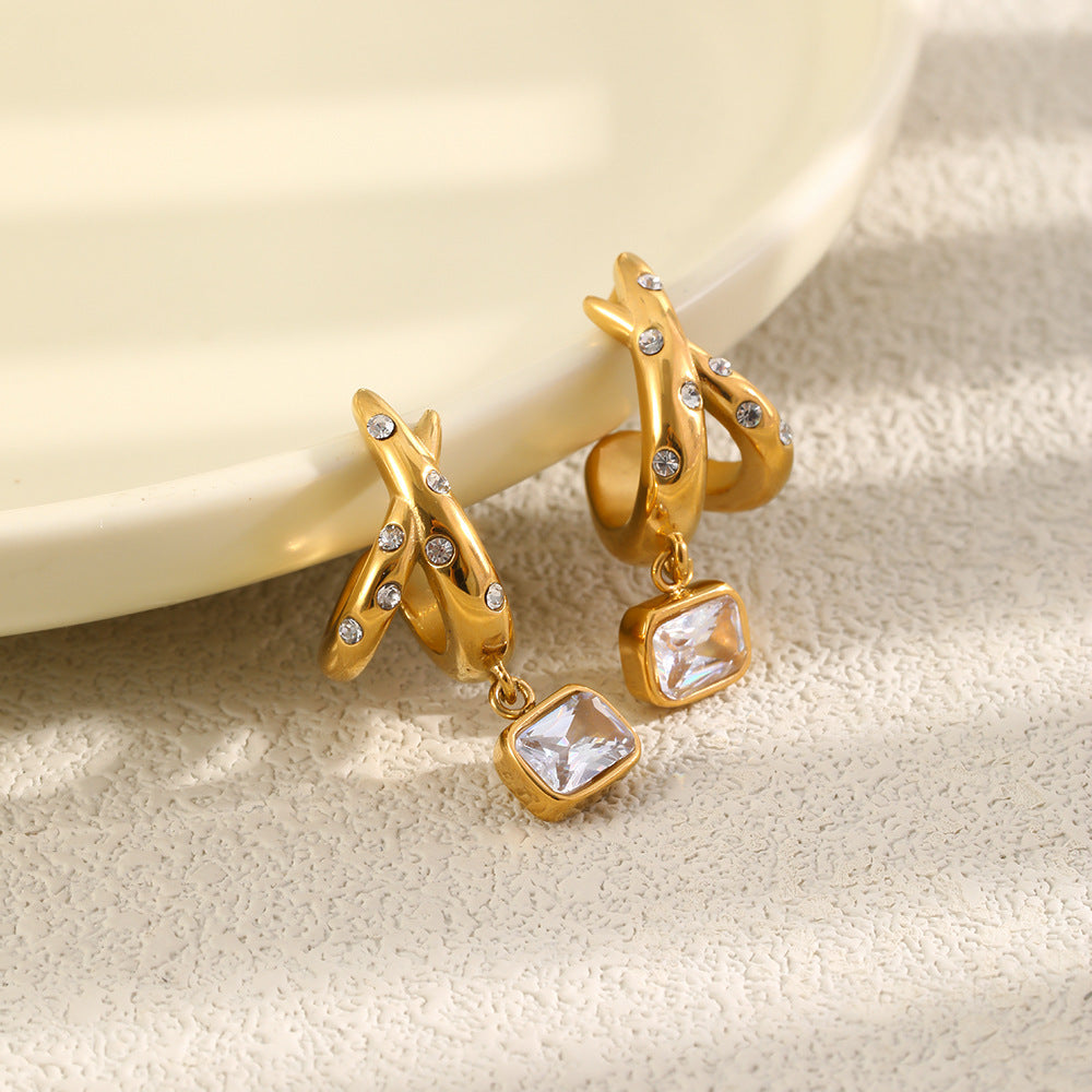 A pair of gold dangle earrings with small clear rhinestones and rectangular clear gemstones, displayed on a textured beige surface next to a cream-colored ceramic plate.