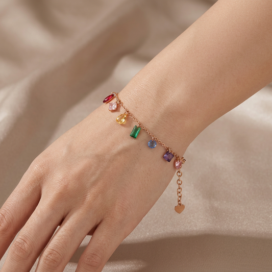 A delicate gold chain bracelet with colorful gemstone charms, shown worn on a person's wrist against a soft beige satin background.