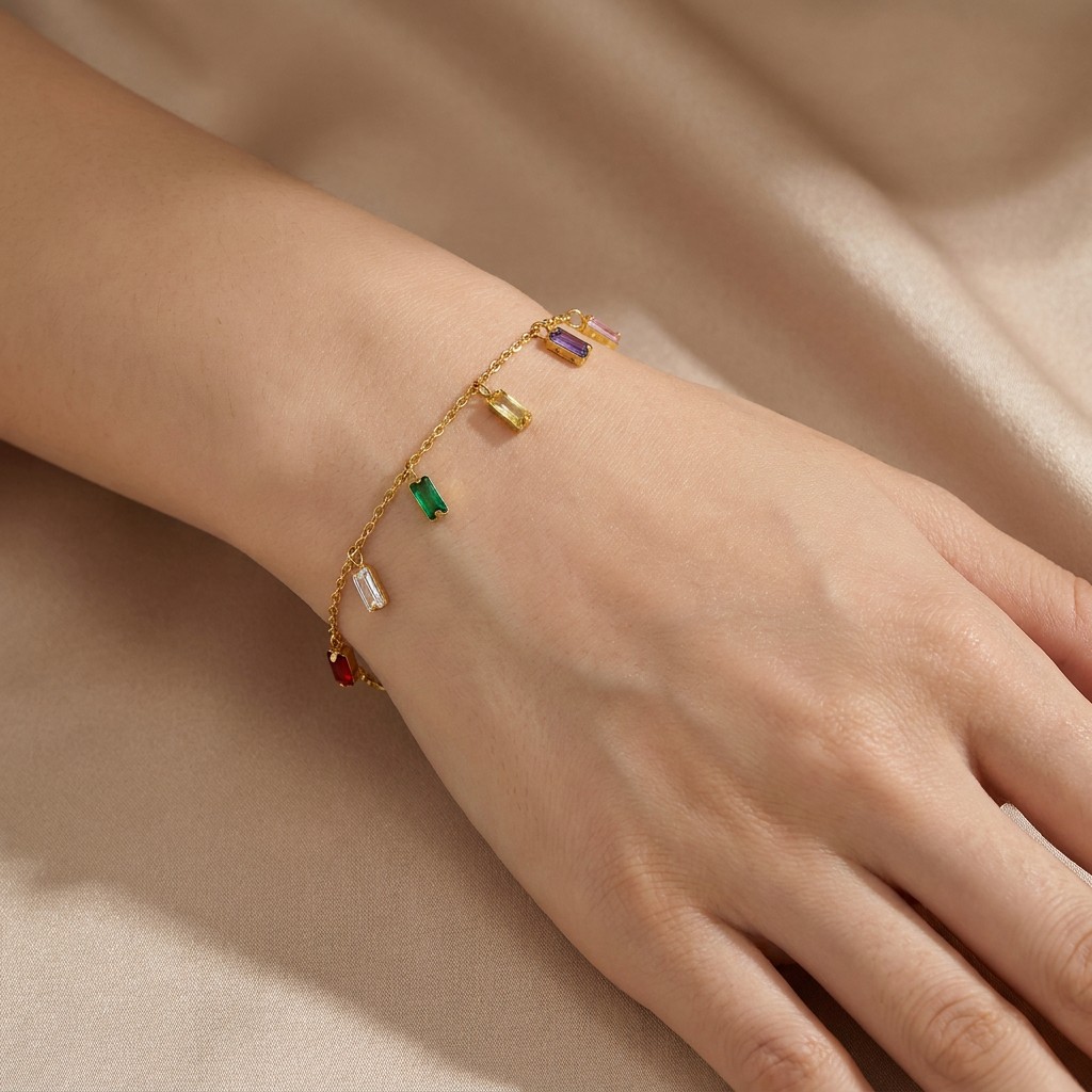 A delicate gold chain bracelet with small colorful rectangular charms, worn on a person's wrist against a soft beige satin background.
