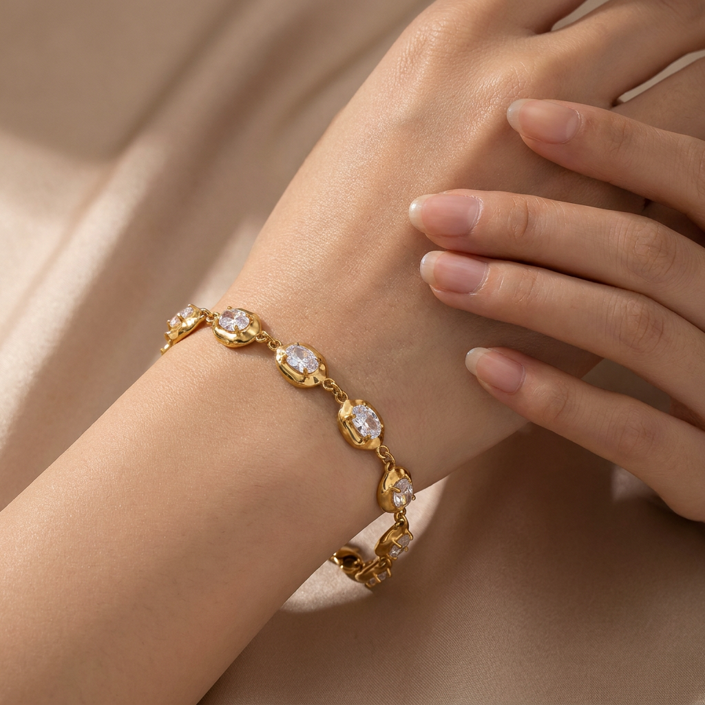 A gold bracelet with clear oval gemstones worn on a person's wrist, shown against a soft beige fabric background.