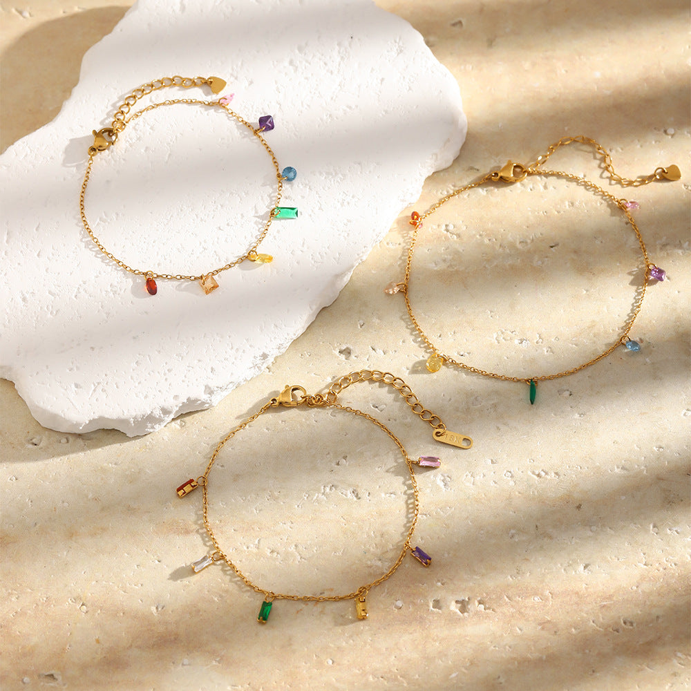 Three delicate gold chain bracelets with small colorful gemstone charms are arranged on a textured beige surface with a white stone prop. The bracelets feature tiny charms in green, purple, red, blue, and gold-tone accents.