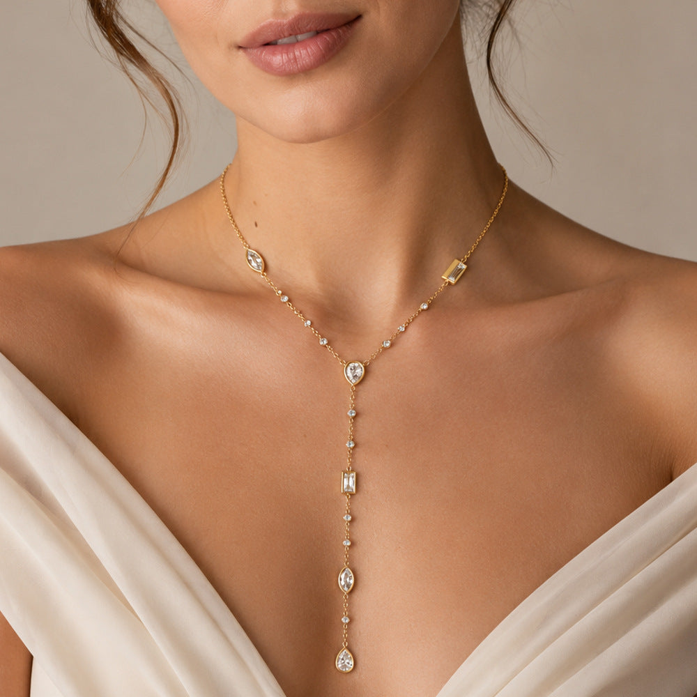 A close-up of a model wearing a delicate gold Y-necklace with clear gemstones, paired with a cream-colored satin dress against a neutral beige background.