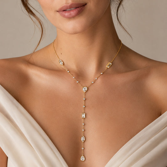 A close-up of a model wearing a delicate gold Y-necklace with clear gemstones, paired with a cream-colored satin dress against a neutral beige background.