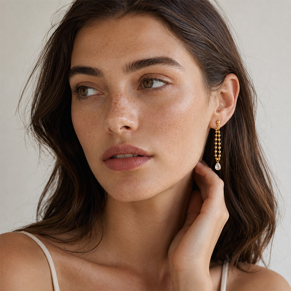 A close-up of a woman wearing a delicate gold beaded drop earring with a teardrop crystal pendant, set against a soft, light beige background.