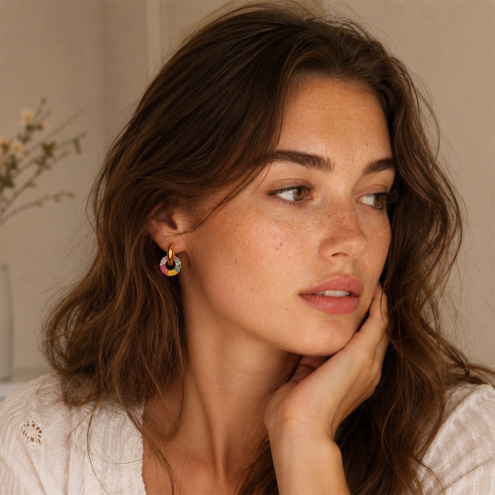 A close-up lifestyle shot of a woman with wavy brown hair wearing a small, colorful rainbow hoop earring. She is resting her chin on her hand, wearing a white textured top against a soft beige background.
