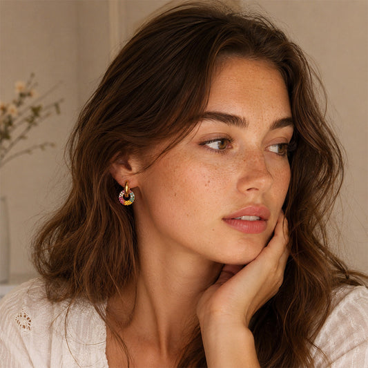 A close-up lifestyle shot of a woman with wavy brown hair wearing a small, colorful rainbow hoop earring. She is resting her chin on her hand, wearing a white textured top against a soft beige background.
