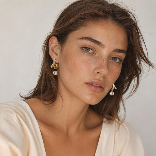 A close-up portrait of a woman wearing gold dangle earrings with clear square-cut stones, against a soft light gray background.