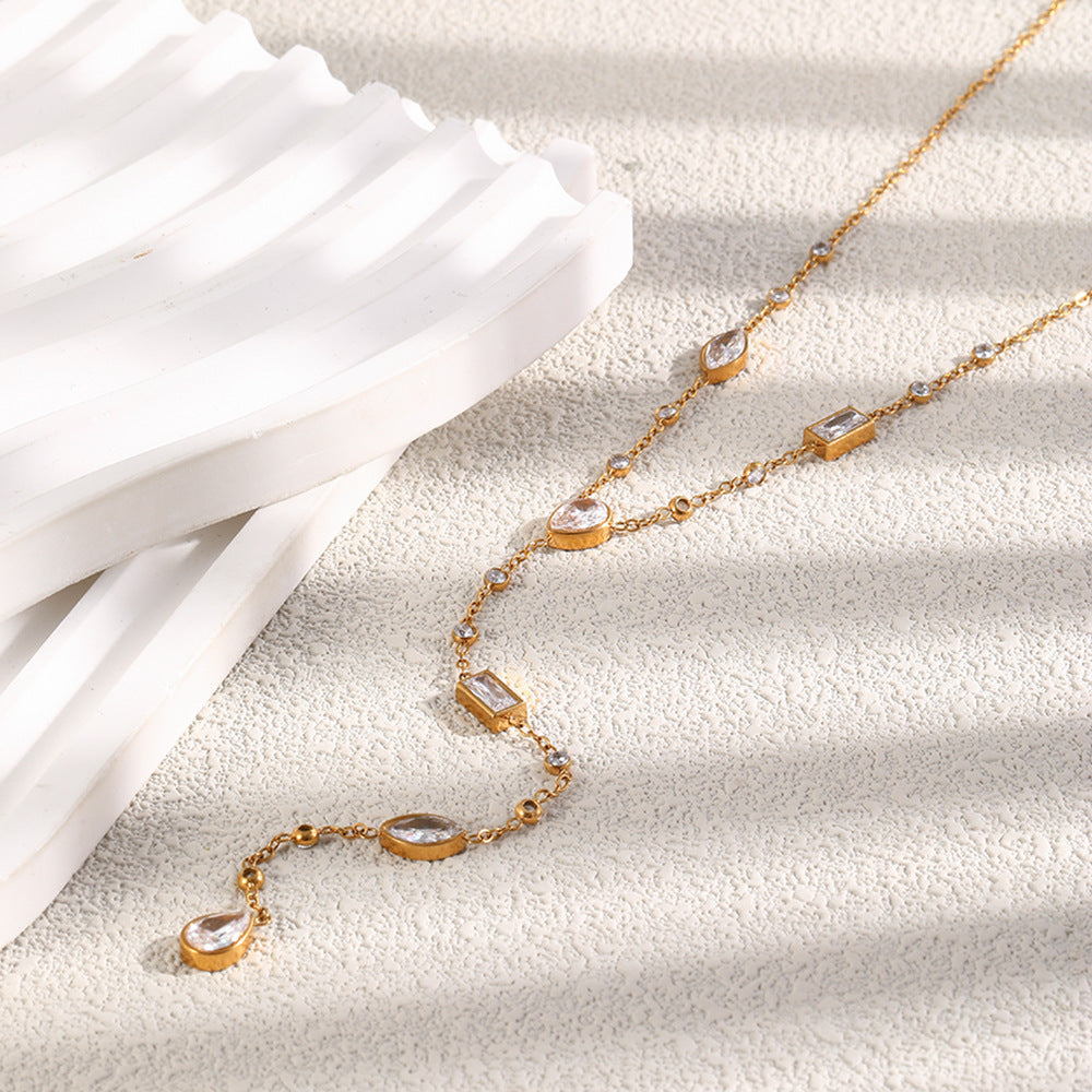 A gold-toned Y-necklace with multiple clear teardrop and rectangular gemstones, styled on a textured white surface next to a white ribbed prop.